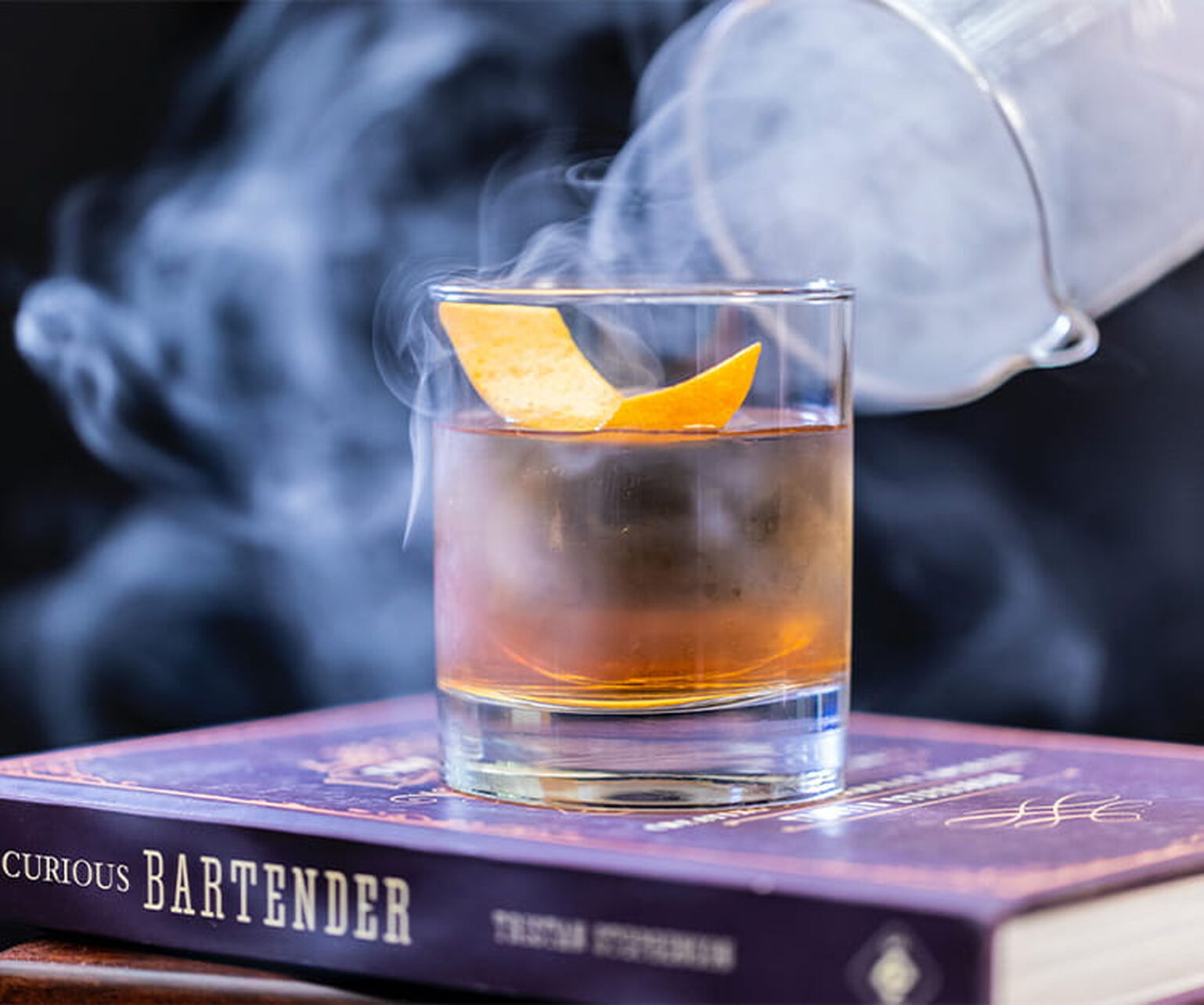 A smoking cloche is taken off an old fashioned cocktail, filling the air with aromatic smoke